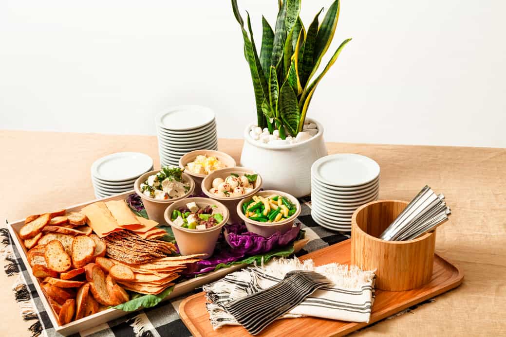 A table with assorted crackers, dips in bowls, stacked plates, cutlery, napkins, and a potted plant.