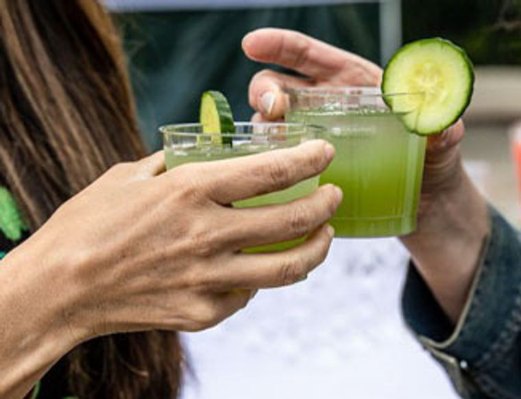 Two people clinking glasses of green cucumber drinks, each garnished with a cucumber slice.