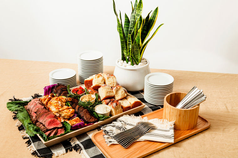 A platter of assorted meats, bread, and dip beside stacked plates, napkins, and a potted plant.
