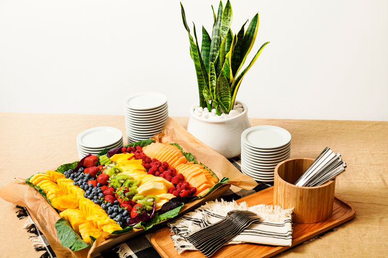 A tray of assorted fresh fruit next to stacks of plates, utensils, and a potted plant on a table.