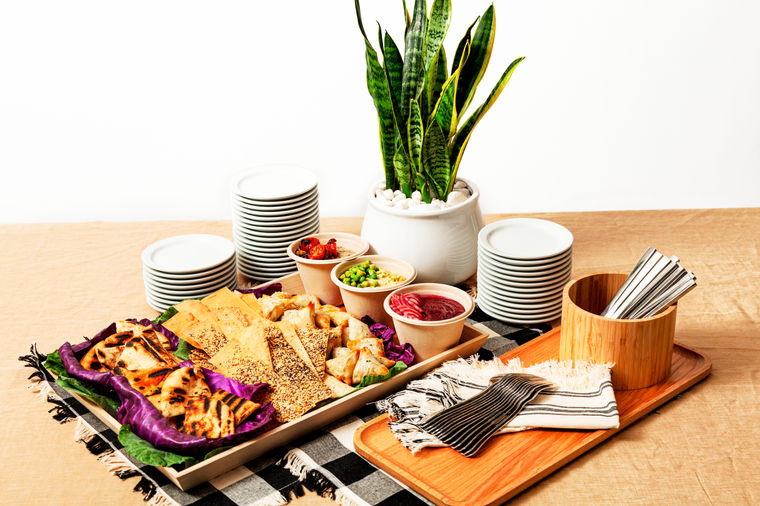 A platter of assorted breads and dips is set on a table with stacked plates, napkins, and a potted plant.