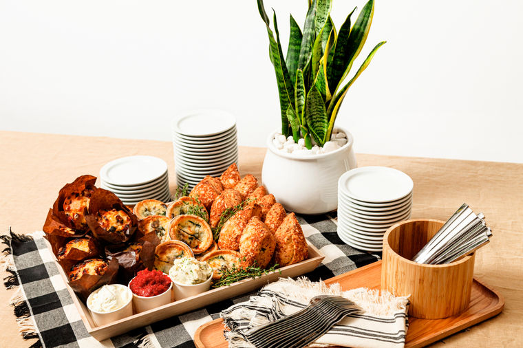A tray of assorted pastries and dips beside stacked plates, napkins, and a potted plant on a checkered cloth.