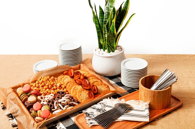 Tray of assorted cookies, pretzels, and macarons beside stacked plates, napkins, and a potted plant on a table.