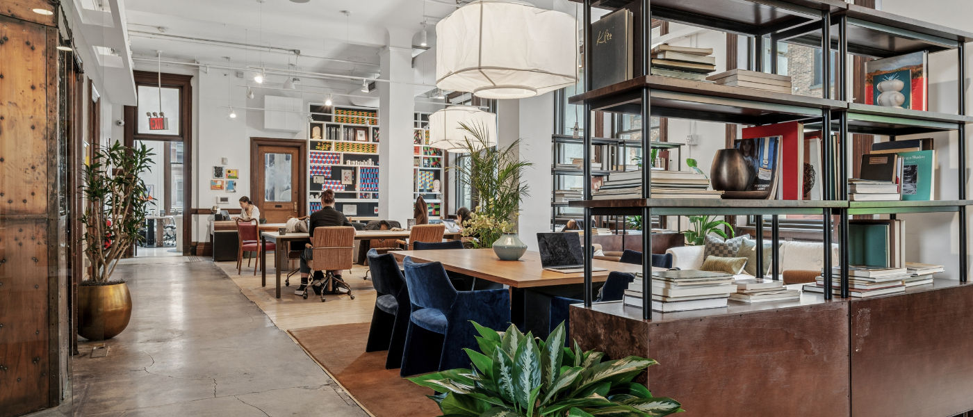 Modern open-plan office with bookshelves, plants, large windows, and people working at shared desks.