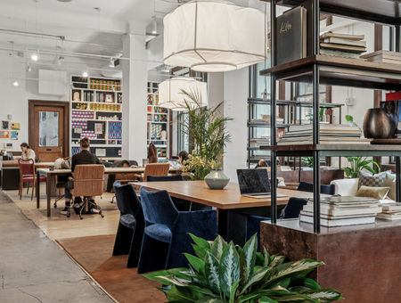 Modern open-plan office with bookshelves, plants, large windows, and people working at shared desks.
