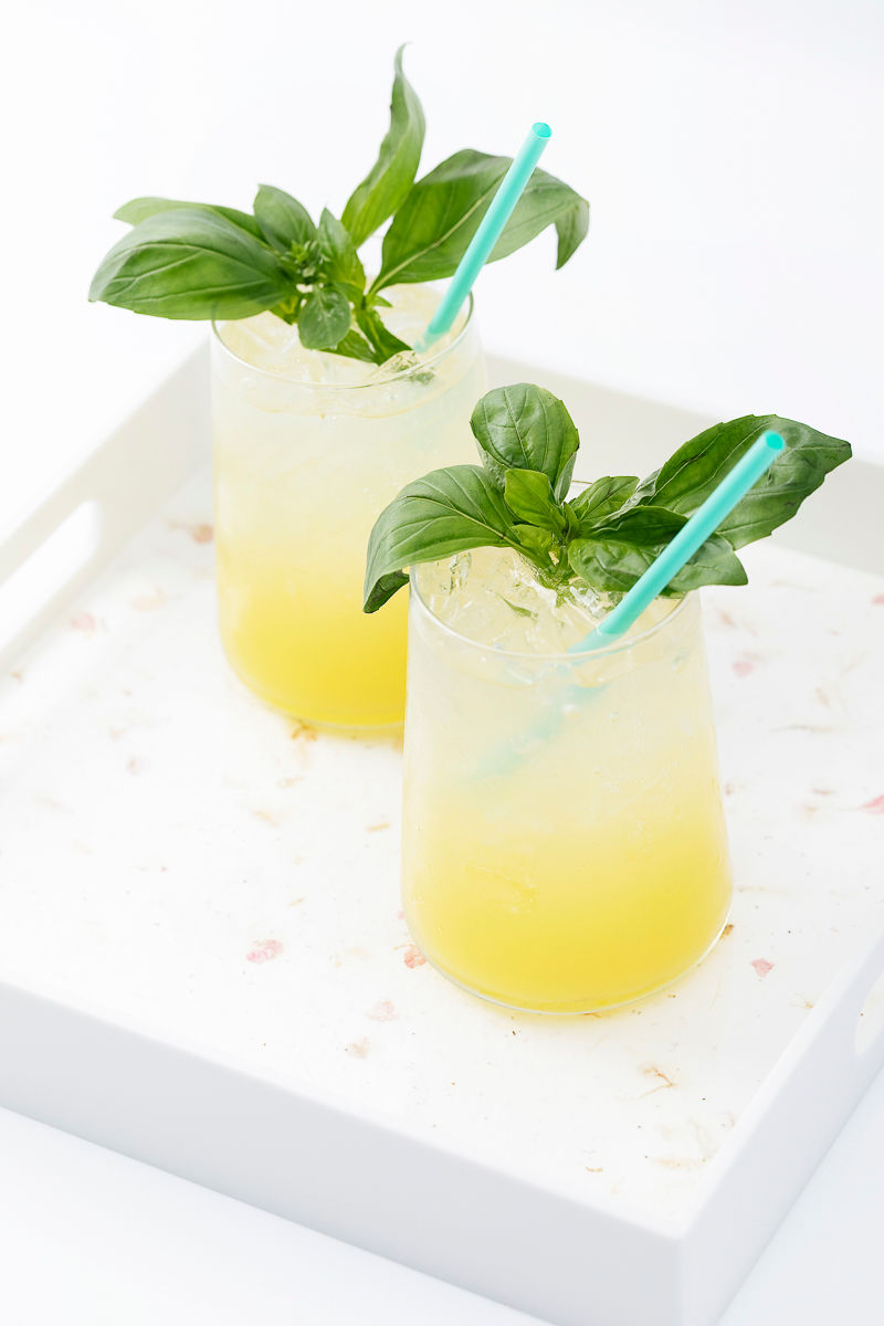 Two yellow cocktails with ice, blue straws, and fresh basil garnish on a white tray.