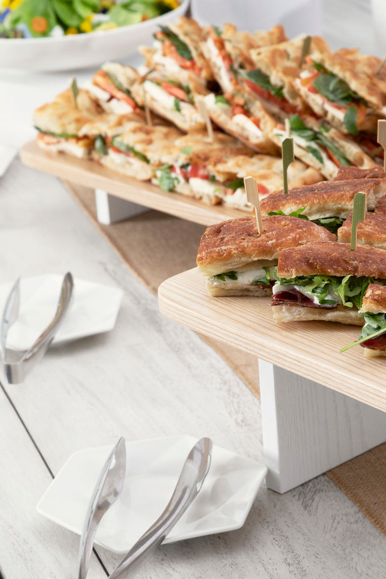Assorted sandwiches with greens on wooden trays, served with tongs and small plates on a light table.
