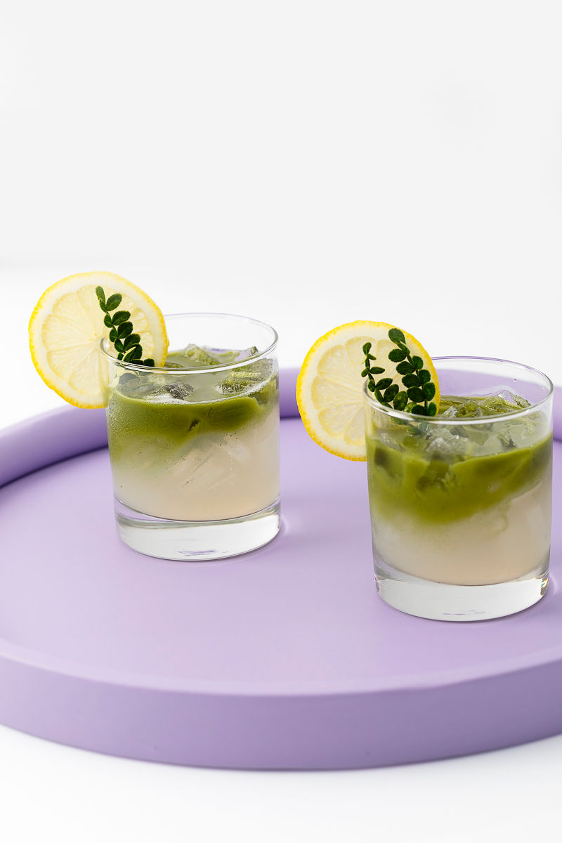 Two iced drinks in clear glasses with lemon slices and greenery, placed on a round lavender tray.