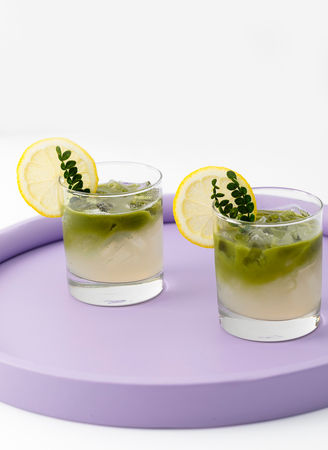 Two iced drinks in clear glasses with lemon slices and greenery, placed on a round lavender tray.