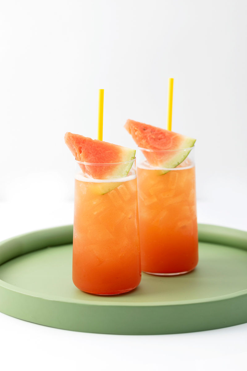 Two iced drinks with orange liquid, garnished with watermelon slices and yellow straws, on a green tray.