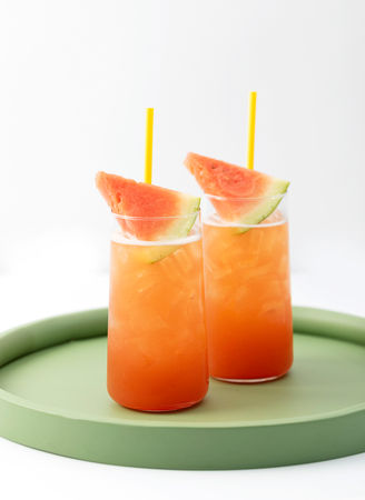 Two iced drinks with orange liquid, garnished with watermelon slices and yellow straws, on a green tray.