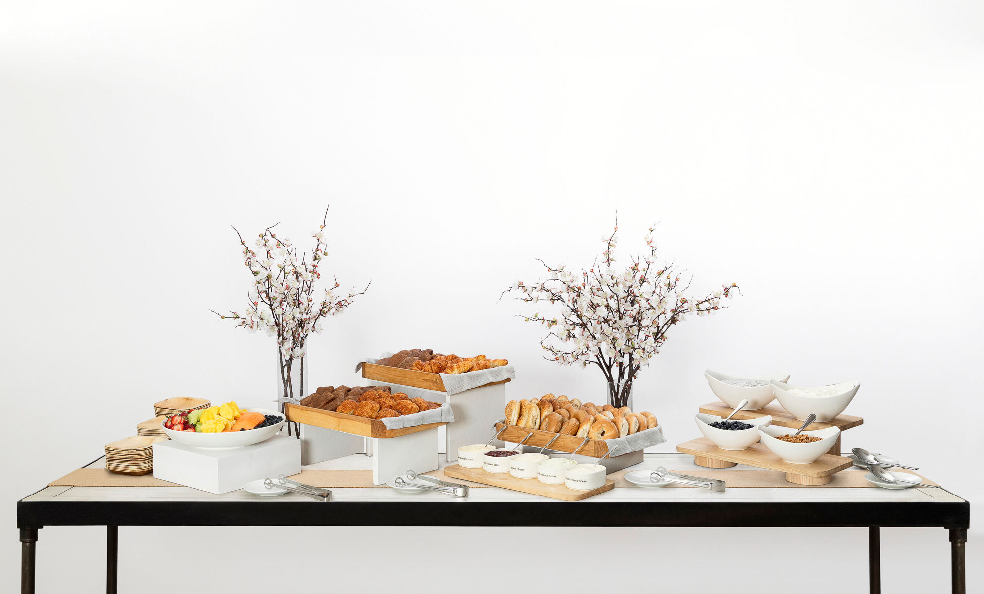 Buffet table with pastries, bagels, fruit, and yogurt, decorated with two vases of white flowering branches.