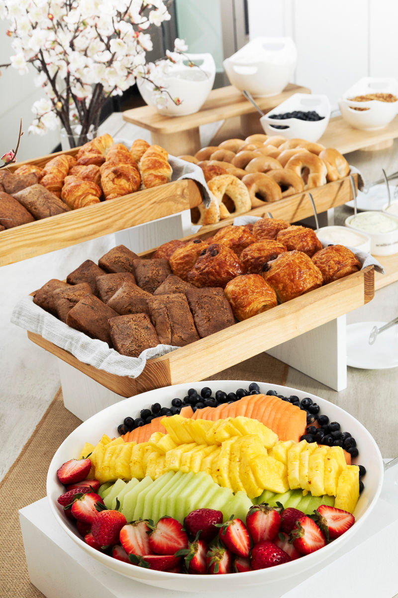 Assorted pastries, bagels, and a fruit platter arranged on buffet tables with white dishes and flowers.