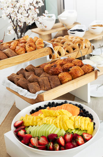 Assorted pastries, bagels, and a fruit platter arranged on buffet tables with white dishes and flowers.