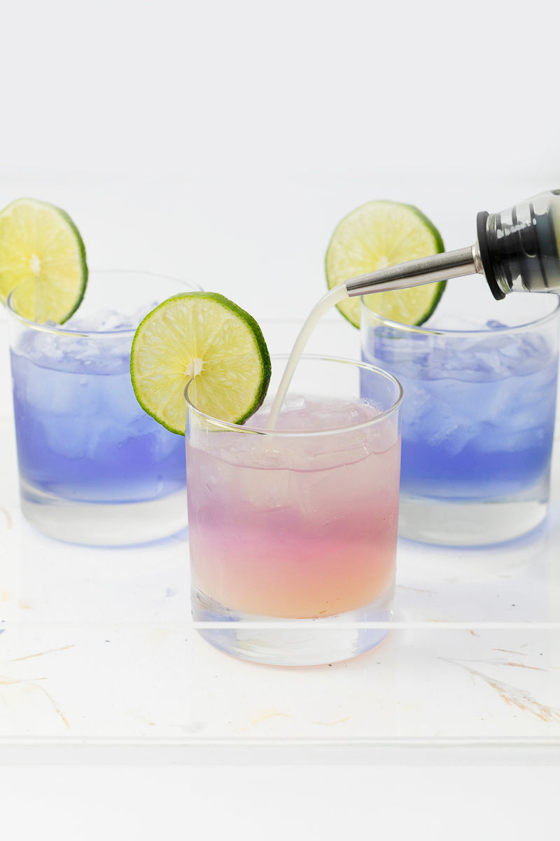 A purple and pink cocktail with a lime slice garnish is being poured, with two blue cocktails in the background.