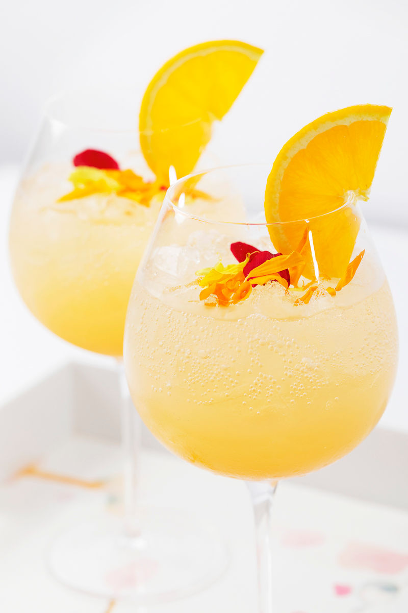 Two glasses of orange cocktails with ice, orange slices, and edible flower petals as garnish.