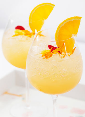 Two glasses of orange cocktails with ice, orange slices, and edible flower petals as garnish.