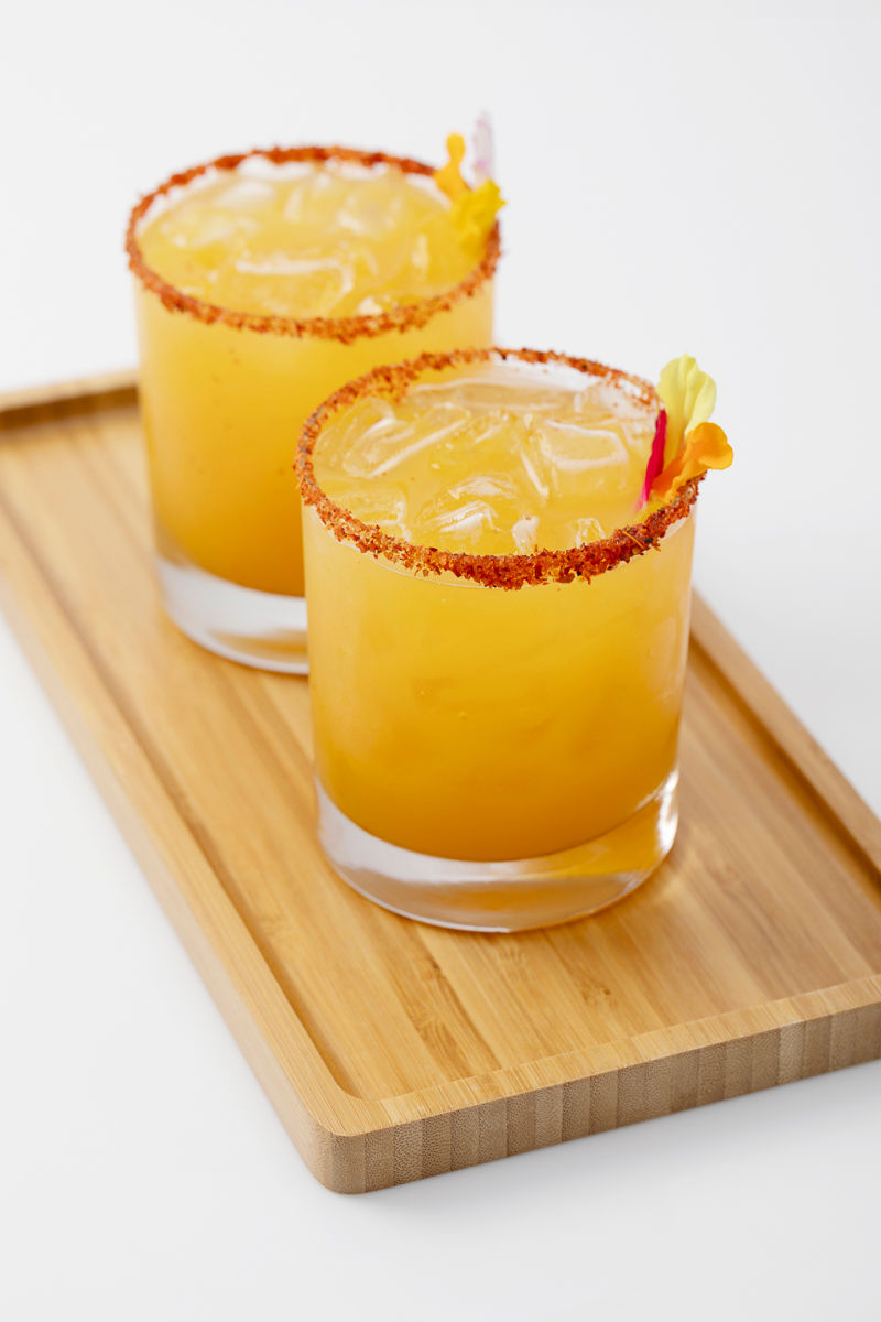 Two orange cocktails with ice, chili rim, and flower garnish on a wooden tray, white background.