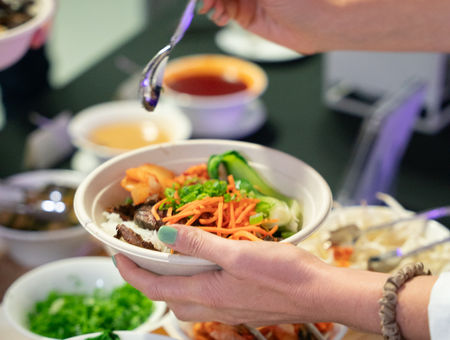 A person adds toppings to a bowl of noodles at a buffet with various vegetables and sauces.
