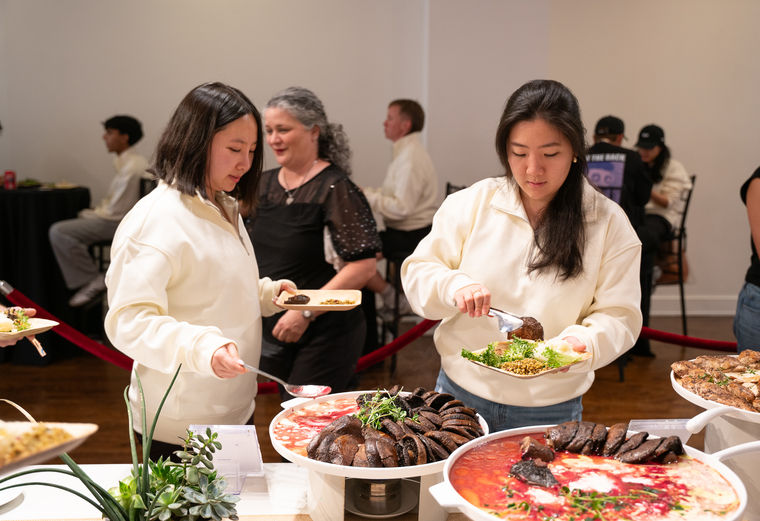 Two women serve food from a buffet table at an indoor event, with others in the background.