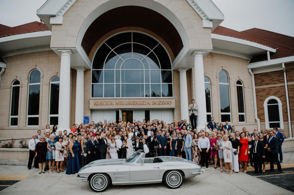 Newlyweds and guests outside church behind vintage car.