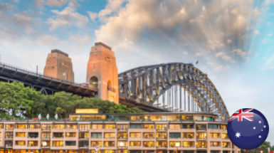 A bridge with towering supports stretches over a waterfront building adorned with an Australian flag in the lower right corner. The sky is partly cloudy with sunlight breaking through, illustrating the scene as if captured by a ผู้เชี่ยวชาญขอวีซ่า traveling the world.