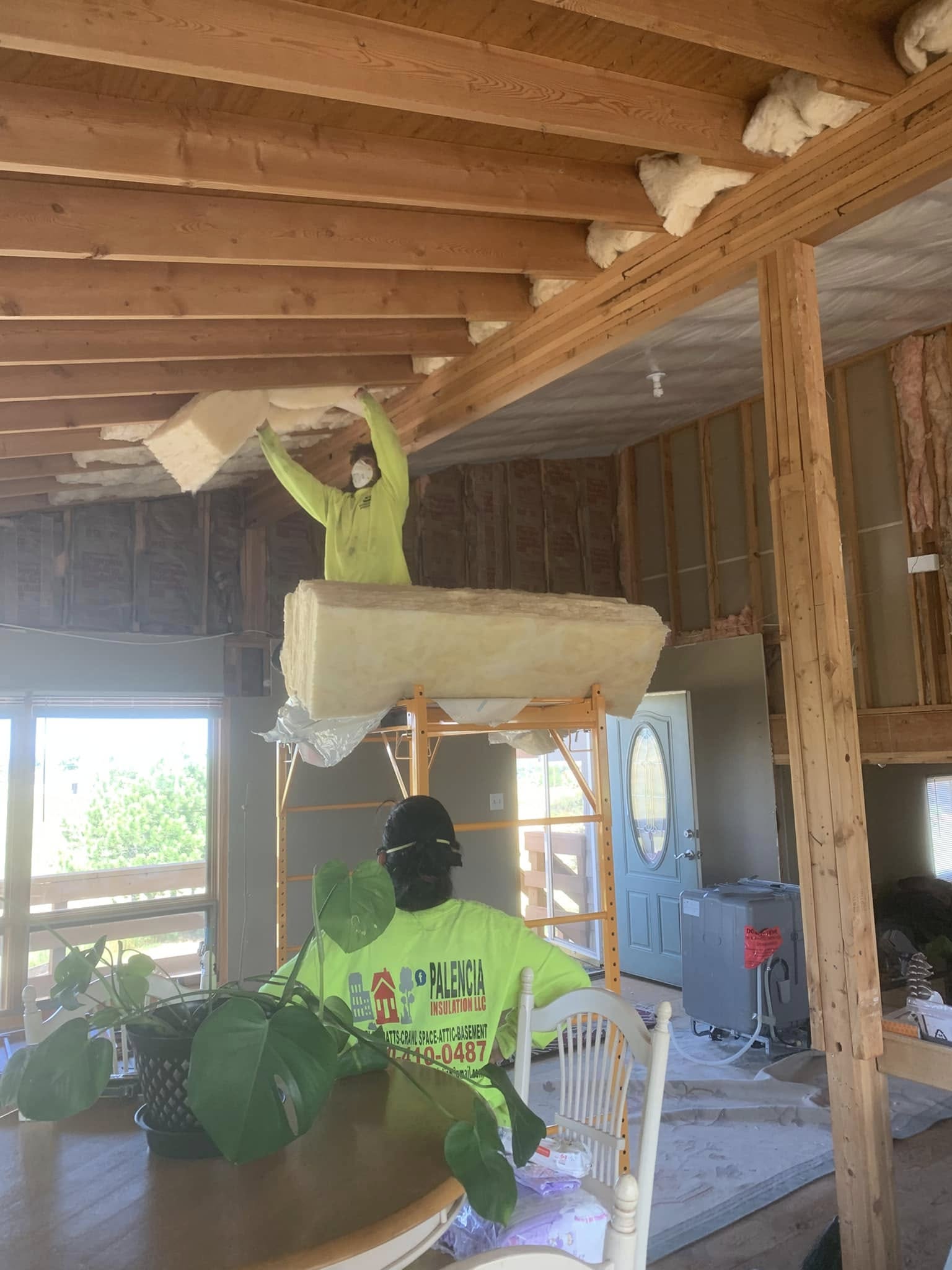 Workers installing fiberglass insulation in a wood-framed vaulted ceiling using scaffolding.