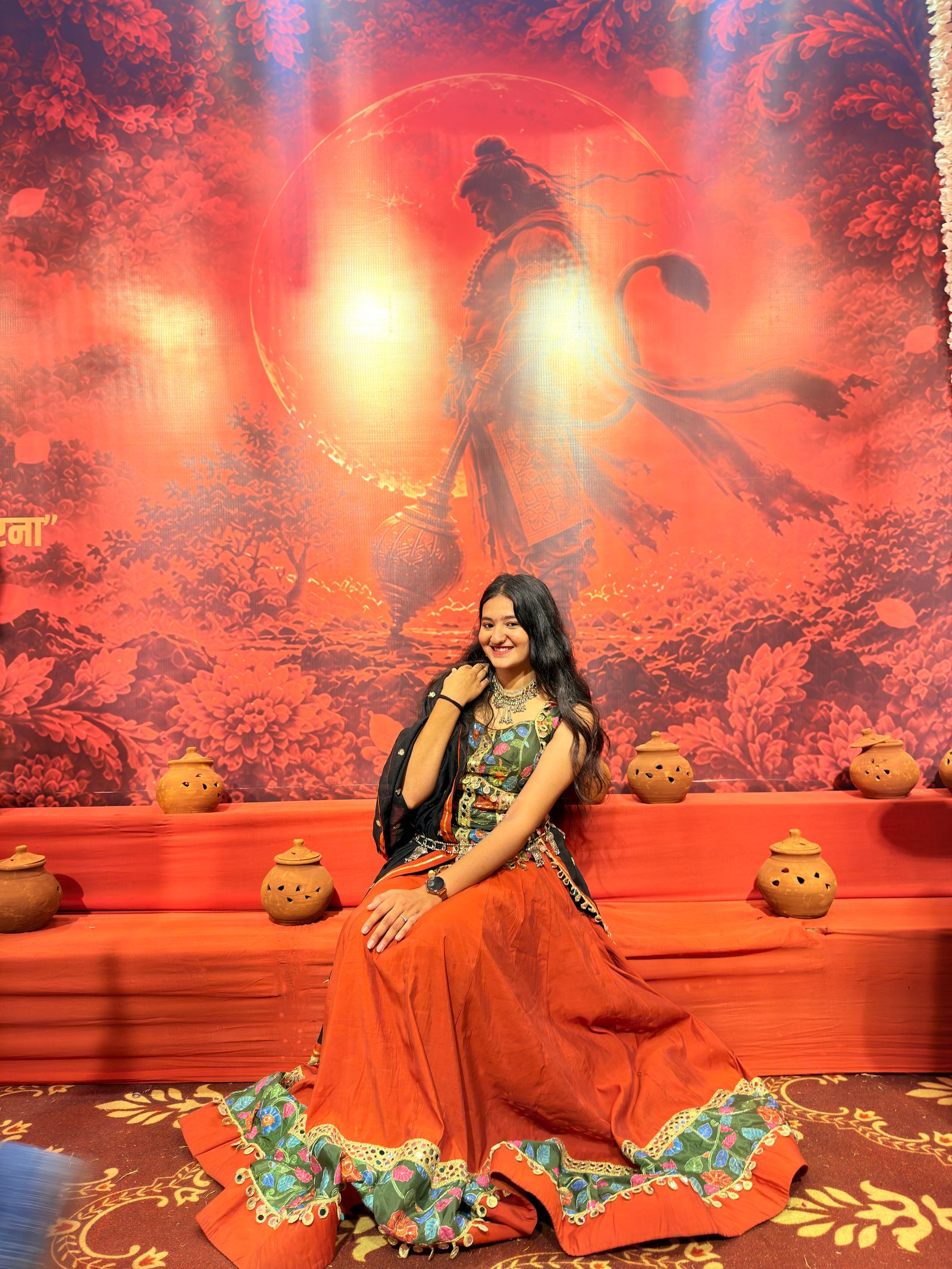 Model seated at a Navratri venue in a rust-red flared chaniya choli with a green floral border, a floral blouse and a black dupatta