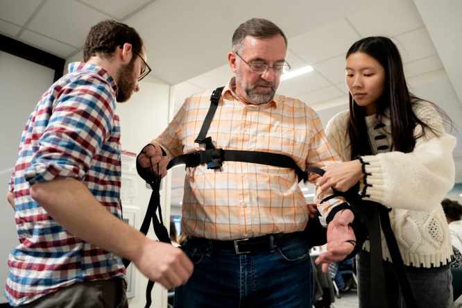 Katie Auyeung, a Washington University junior engineering major from Chicago, right, and Zachary Fine, a sophomore biomedical engineering major from Silver Spring, Md., left, help Craig Oberle, of Florissant, put on a device they invented that would help limit his hand tremors at the end of the school’s annual Make-a-Thon program on Thursday in St. Louis’ Central West End neighborhood. (Brian Munoz / St. Louis Public Radio)