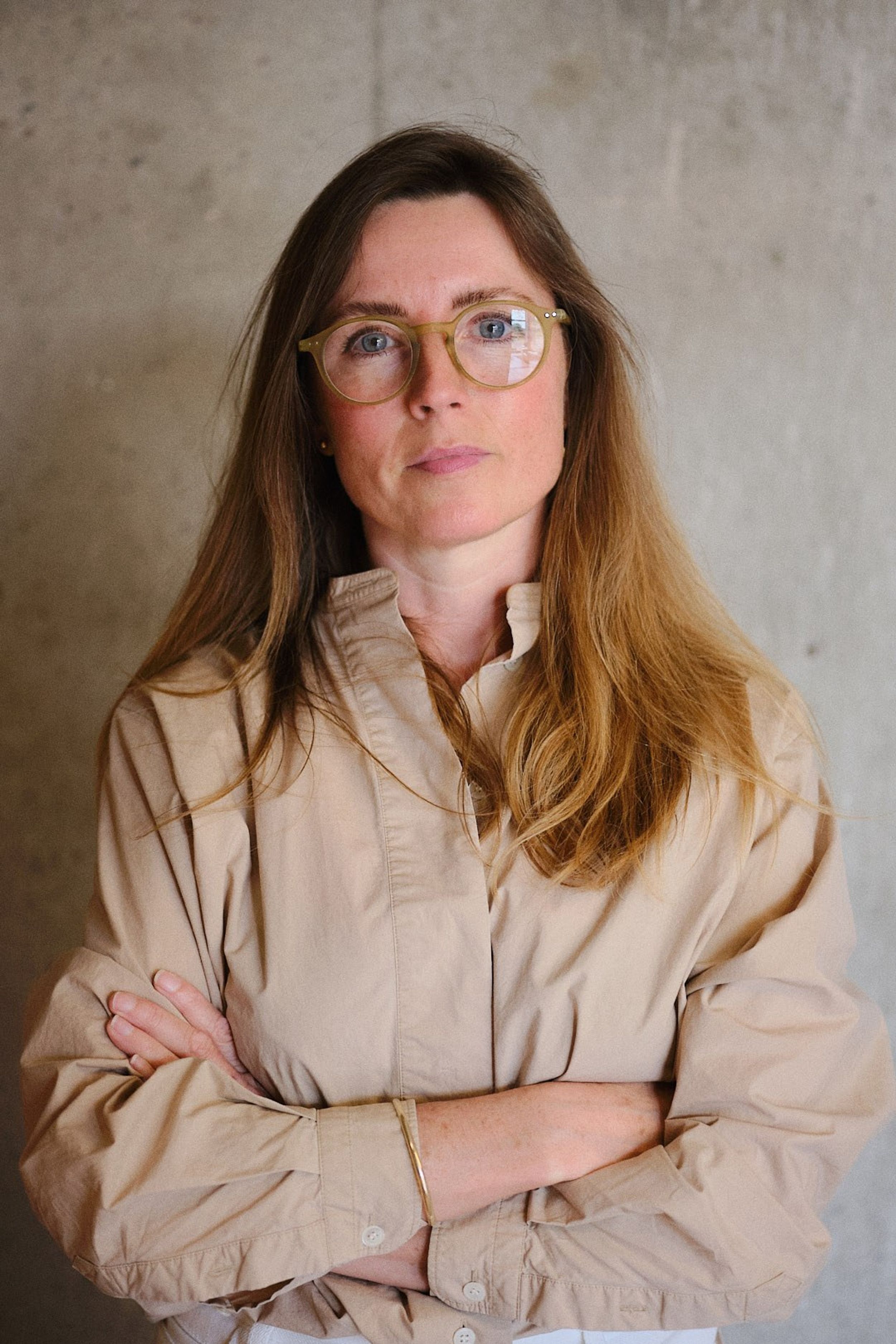 A profile shot of a woman with glasses and brown hair wearing a beige shirt and crossed arms.