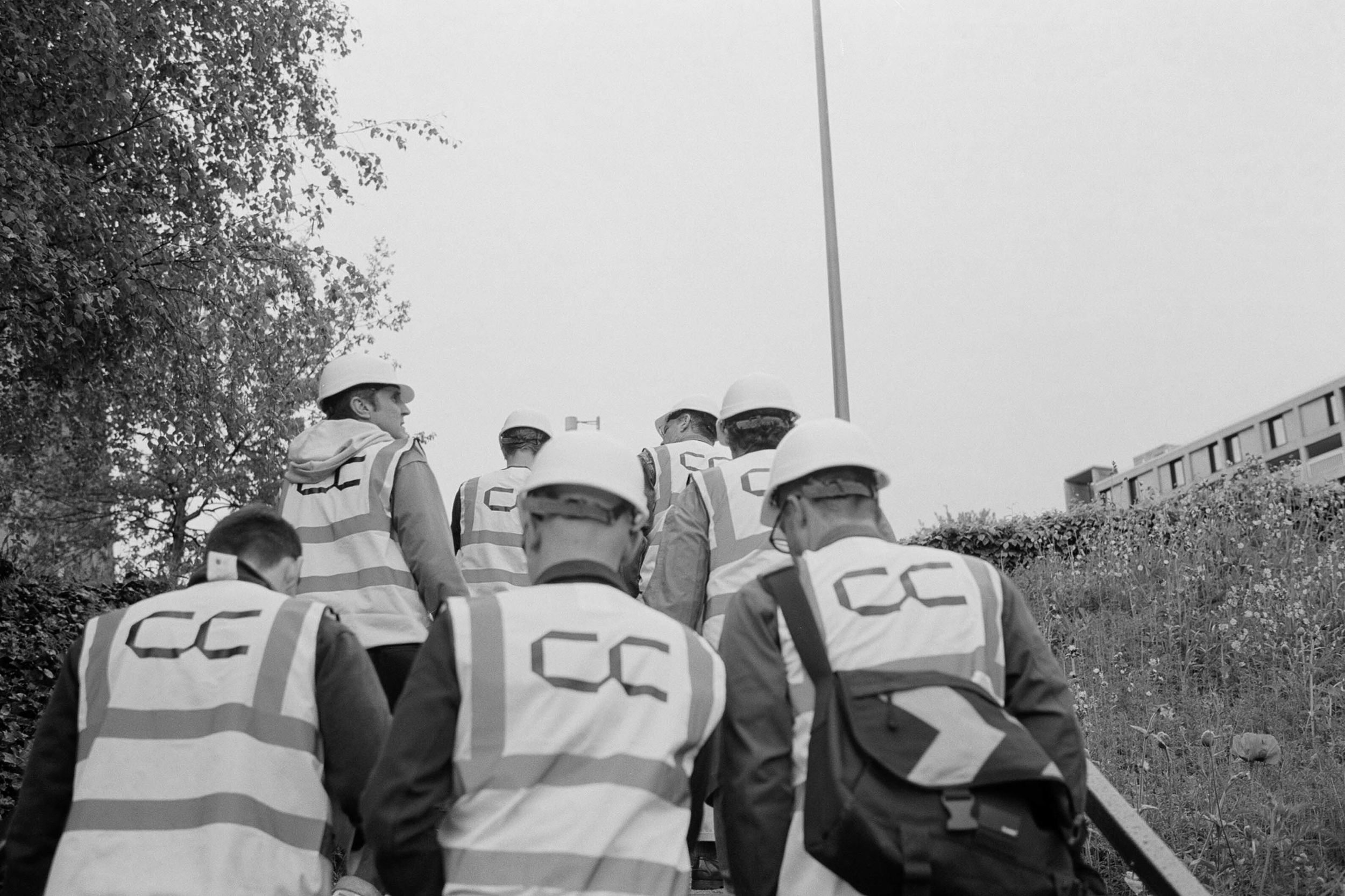 Shot from behind, a group of people wearing hard hats and hi-vis with Concrete Communities logo printed on the back of the jacket.