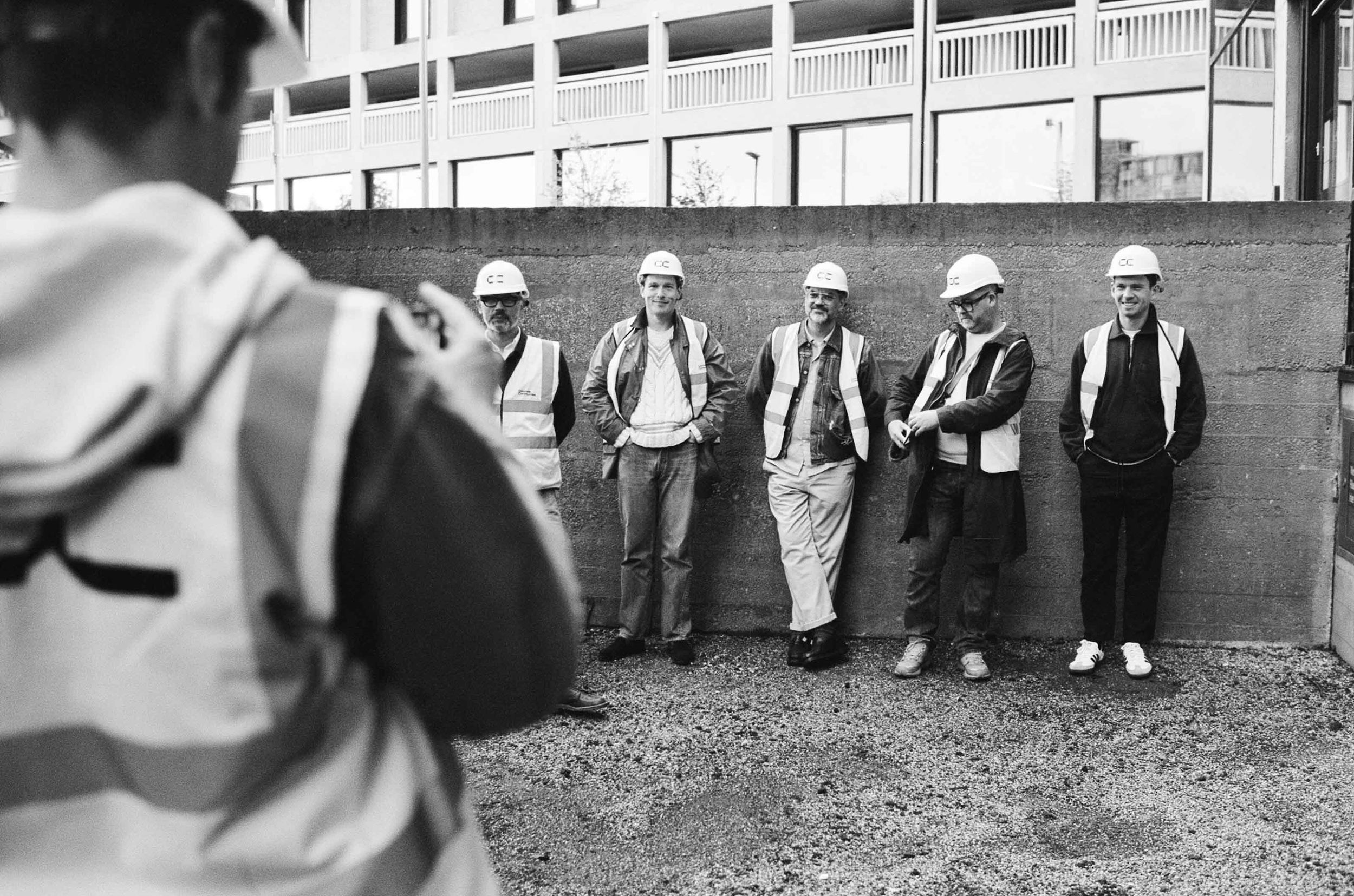 A group of five men in hi-vis and hard hats standing against a concrete wall having their photo taken.