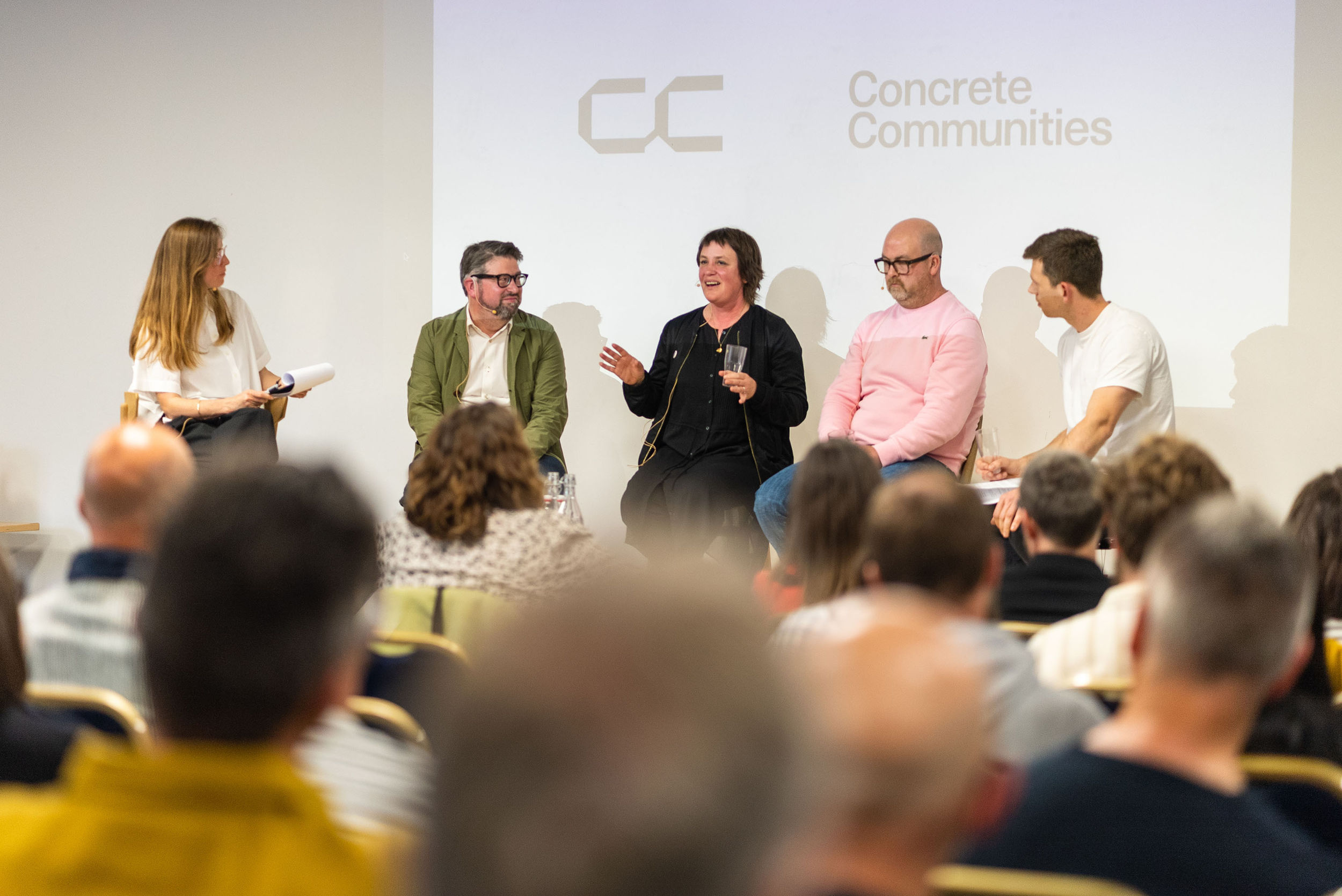 A panel of five people debate on a stage with audience in foreground.