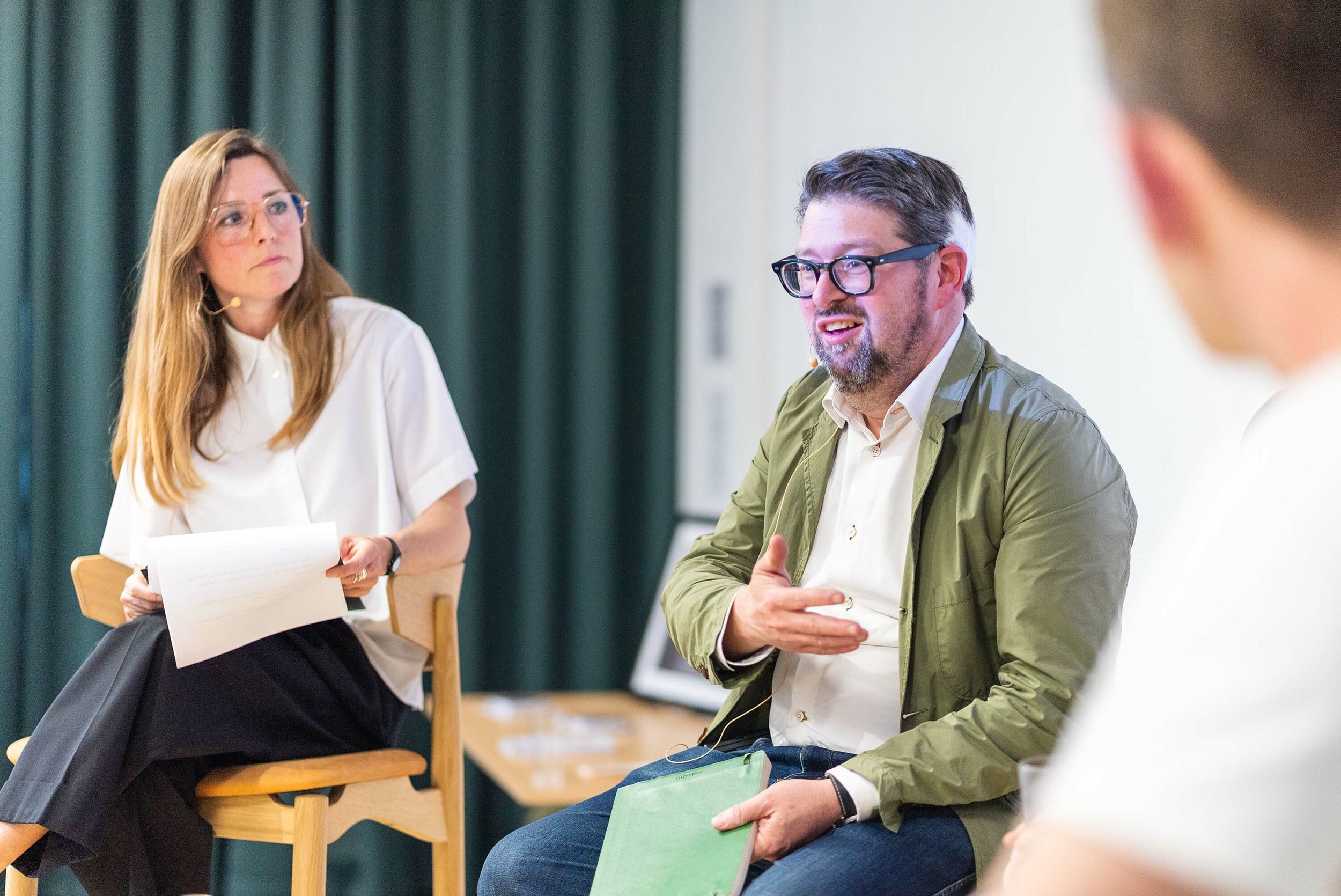 A panel of people having a debate with the focus on a woman in a white shirt and glasses and a man in a green jacket and glasses.