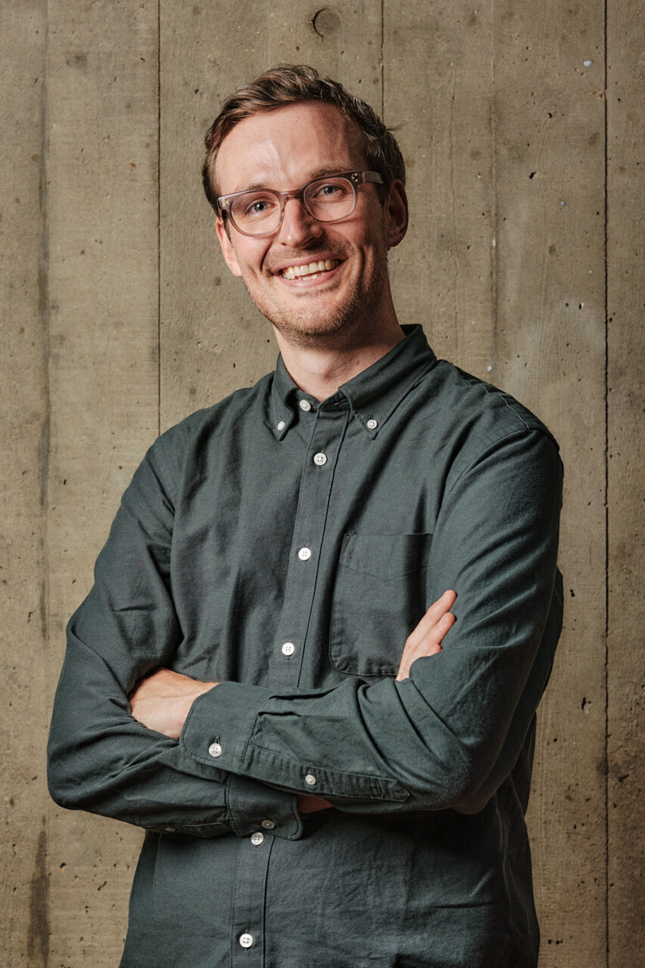 A profile shot of a man smiling with crossed arms wearing glasses and a green shirt, in front of a board marked concrete backdrop.