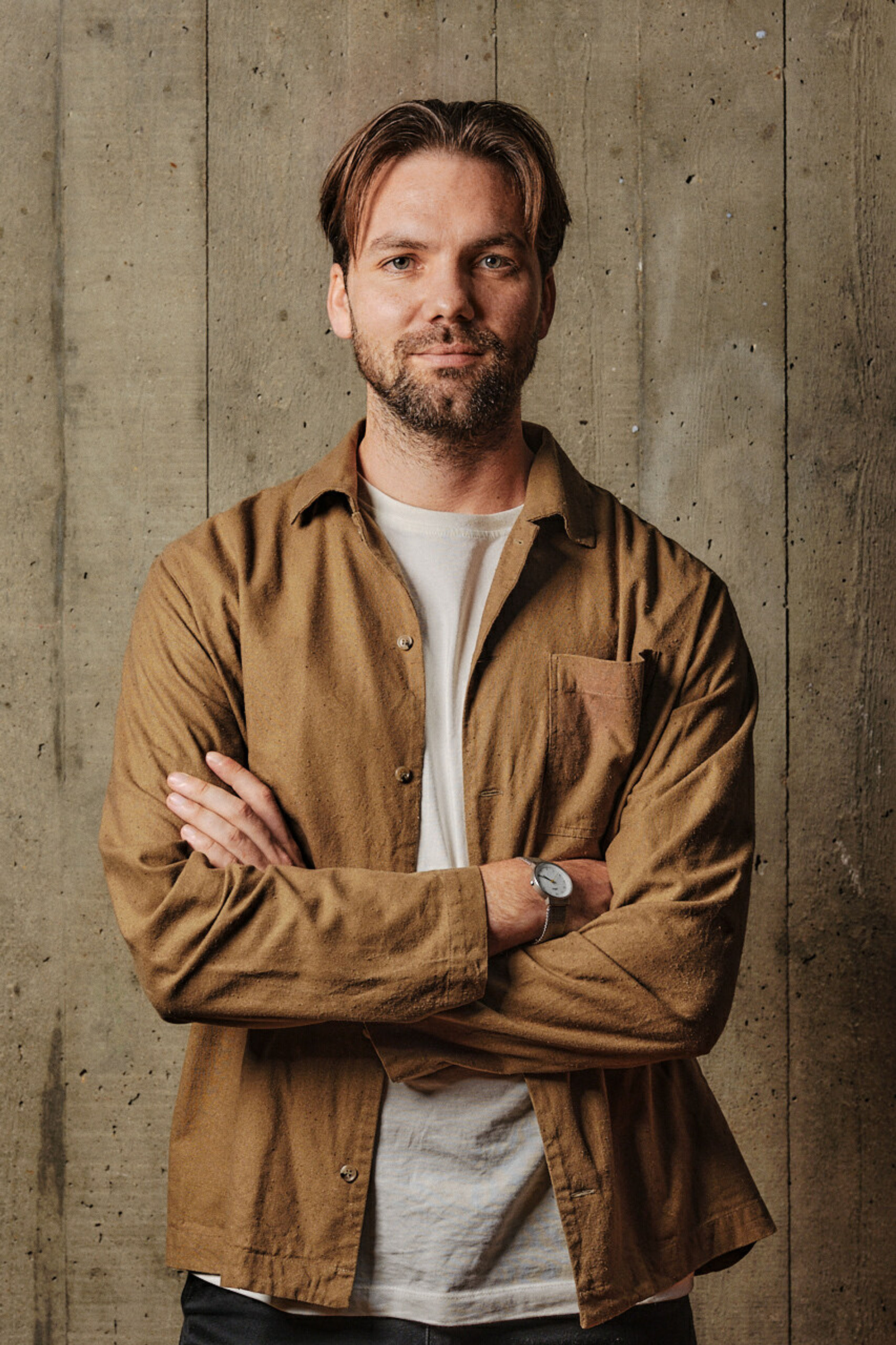 A profile shot of a man with crossed arms wearing a brown overshirt, white t-shirt and watch, in front of a board marked concrete backdrop.