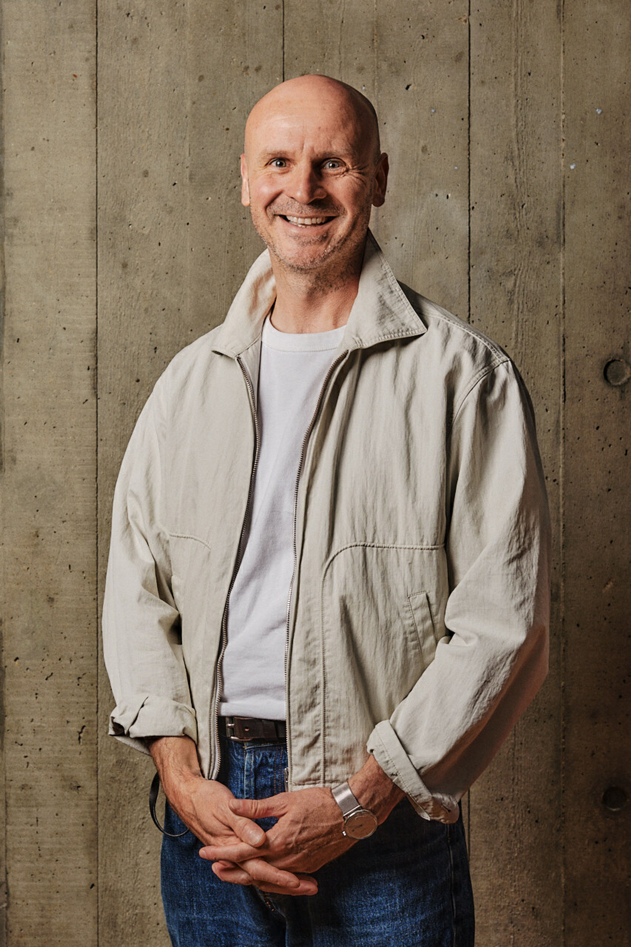 A profile shot of a smiling man, wearing a beige open shirt and white t-shirt, in front of a board marked concrete backdrop.