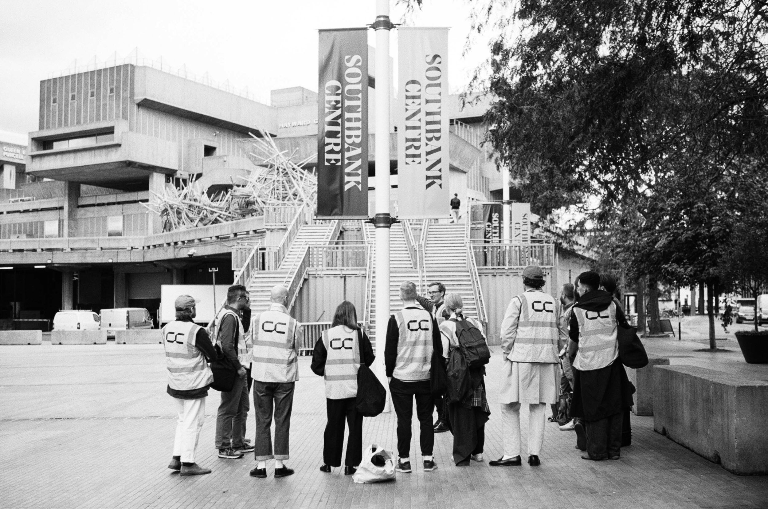 Shot from behind, a tour guide and group of people wearing hi-vis with Concrete Communities logo printed on the back at the Southbank Centre, London.