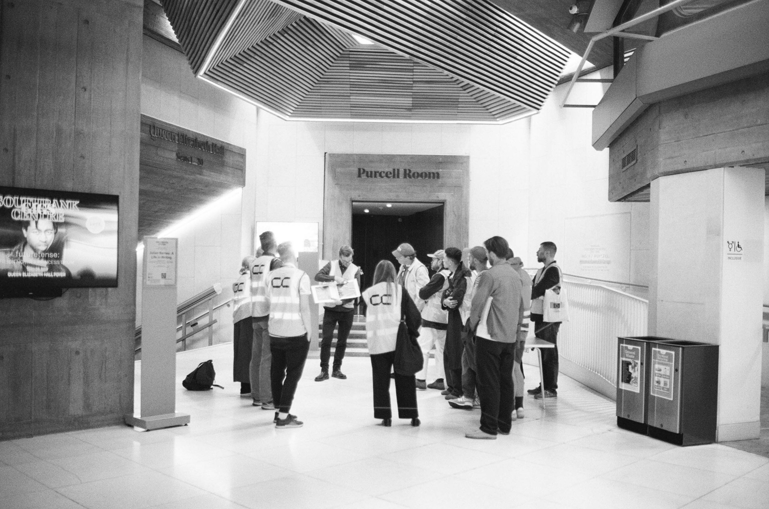 Shot from behind, a group of people wearing hi-vis with Concrete Communities logo printed on the back at the Purcell Room in Southbank Centre, London.