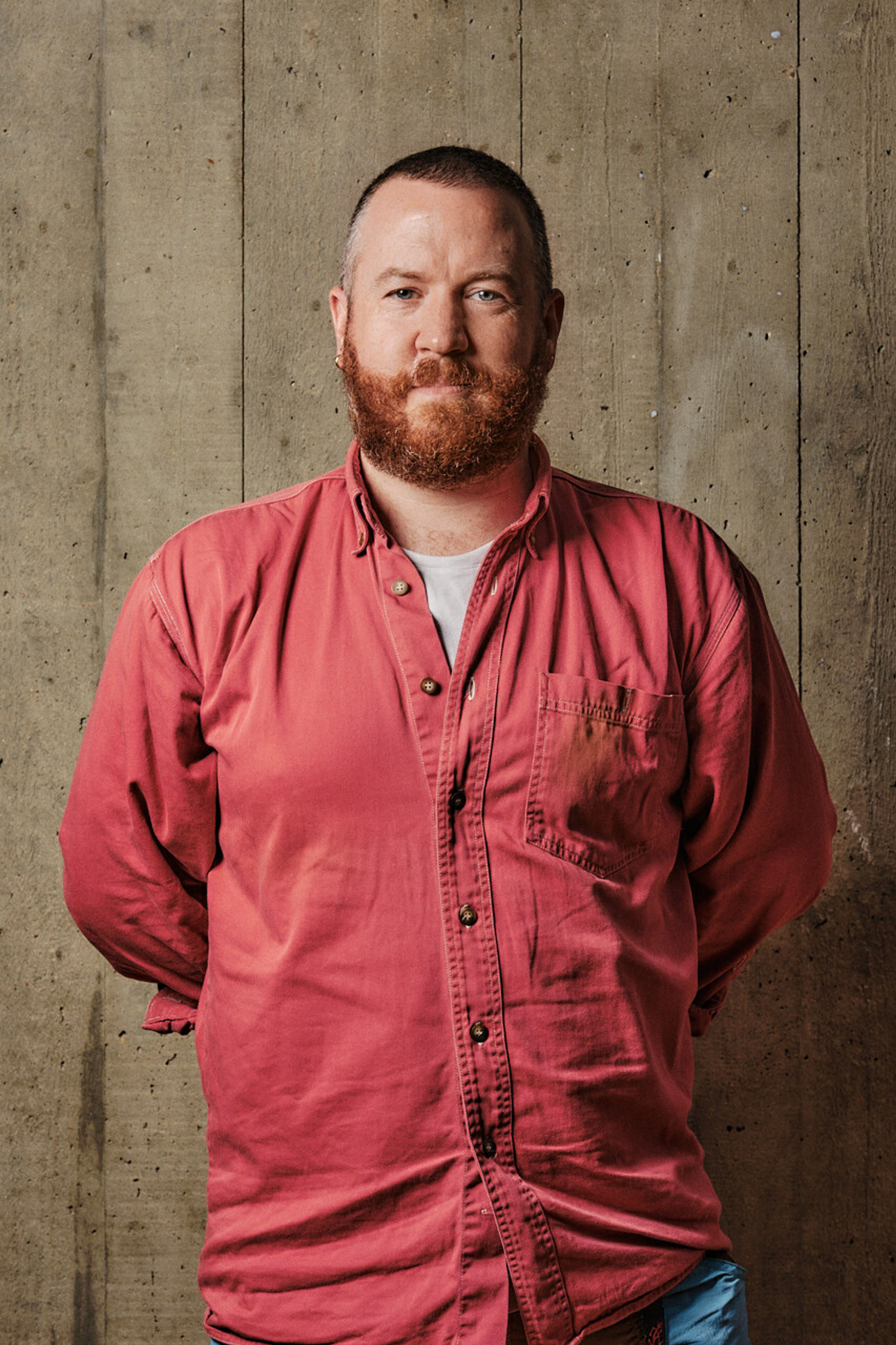 A profile shot of a smiling man with facial hair, with wearing a red shirt, in front of a board marked concrete backdrop.