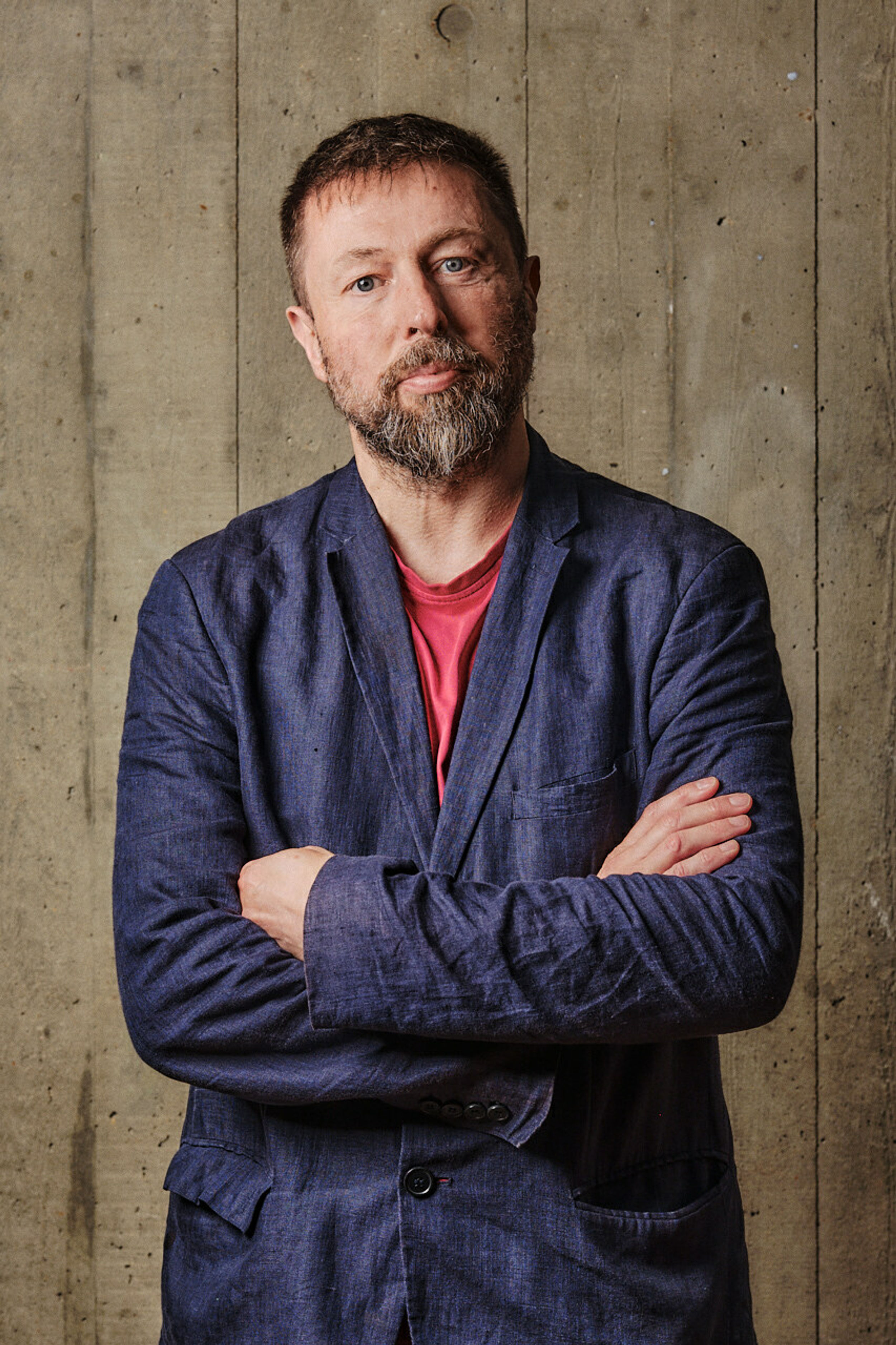 A profile shot of a man with facial hair with crossed arms wearing a denim jacket, in front of a board marked concrete backdrop.