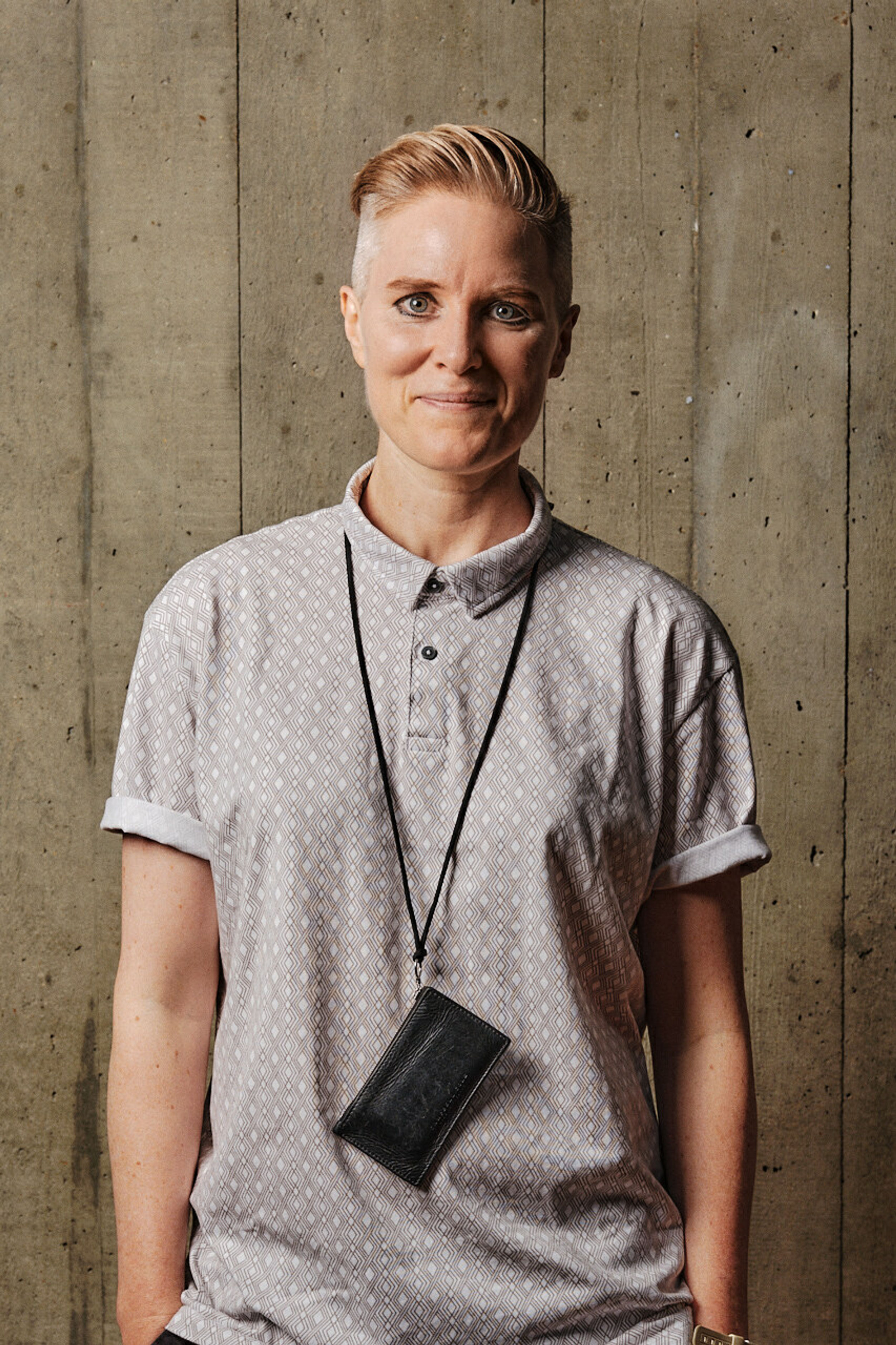 A profile shot of a person smiling with short blonde hair, wearing a patterned, short sleeved shirt and black wallet around their neck, in front of a board marked concrete backdrop.