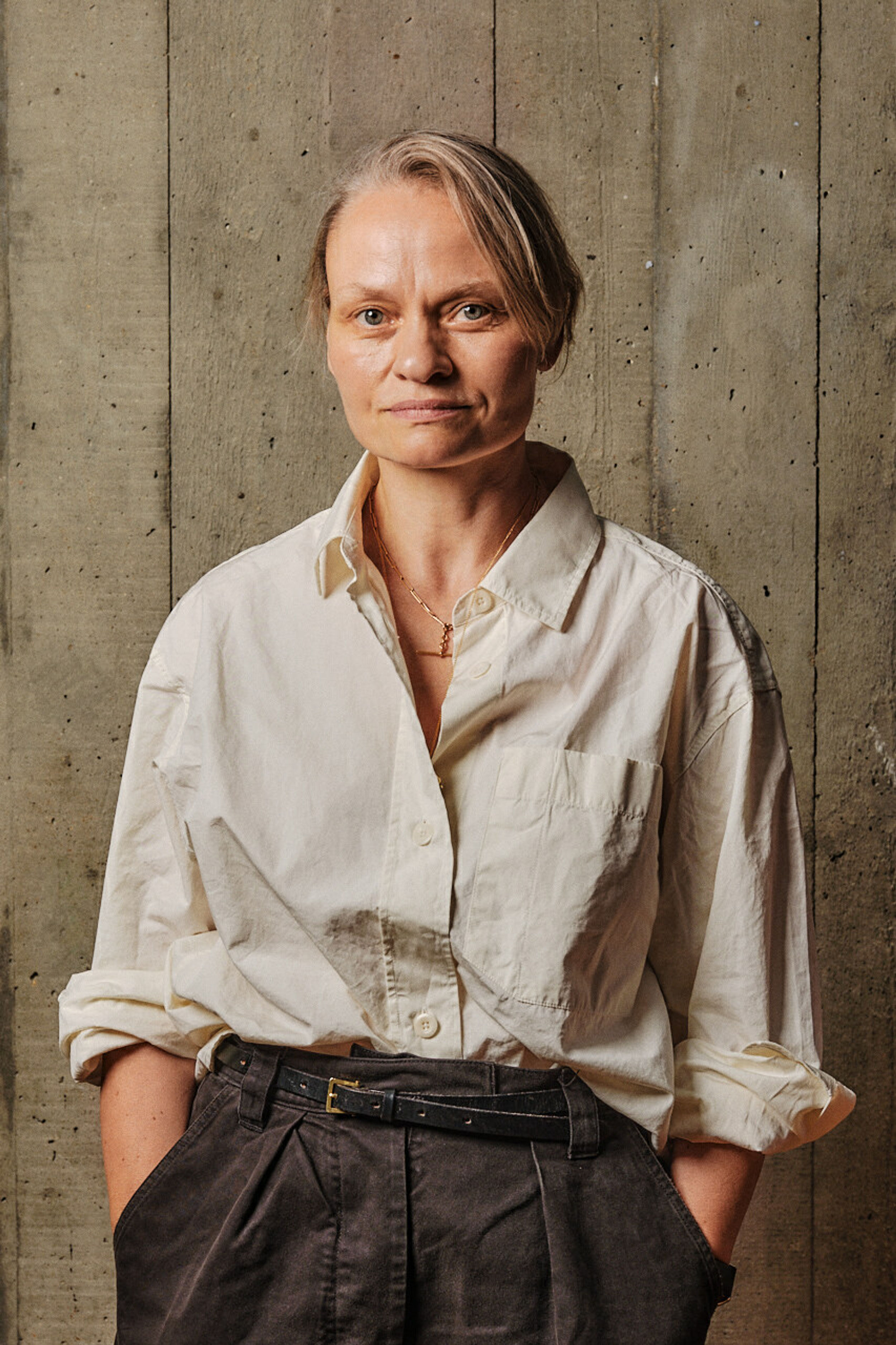 A profile shot of a woman with light hair wearing an open collared shirt, in front of a board marked concrete backdrop.