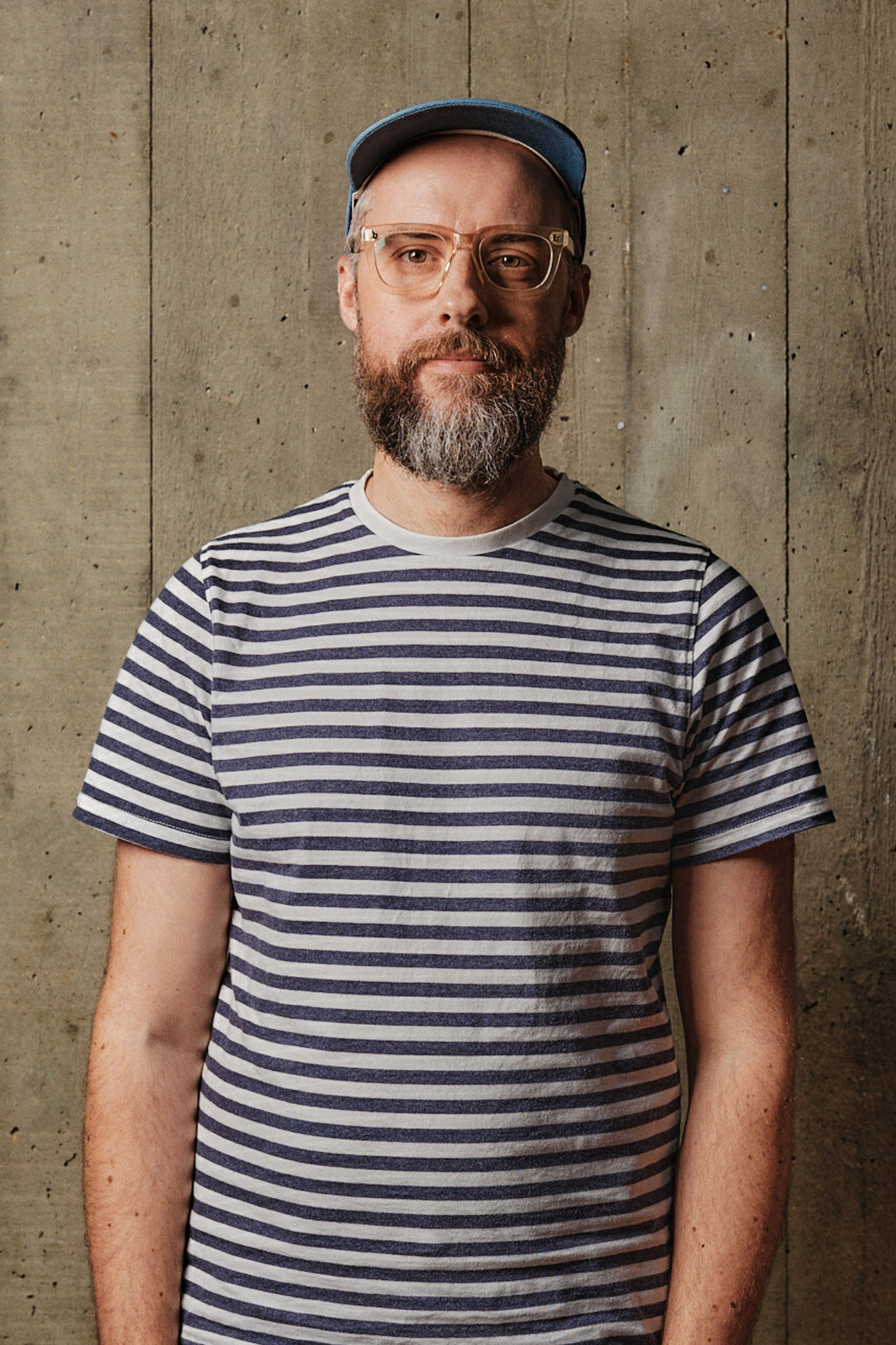 A profile shot of a smiling man with facial hair, wearing glasses, a striped t-shirt and cap, in front of a board marked concrete backdrop.