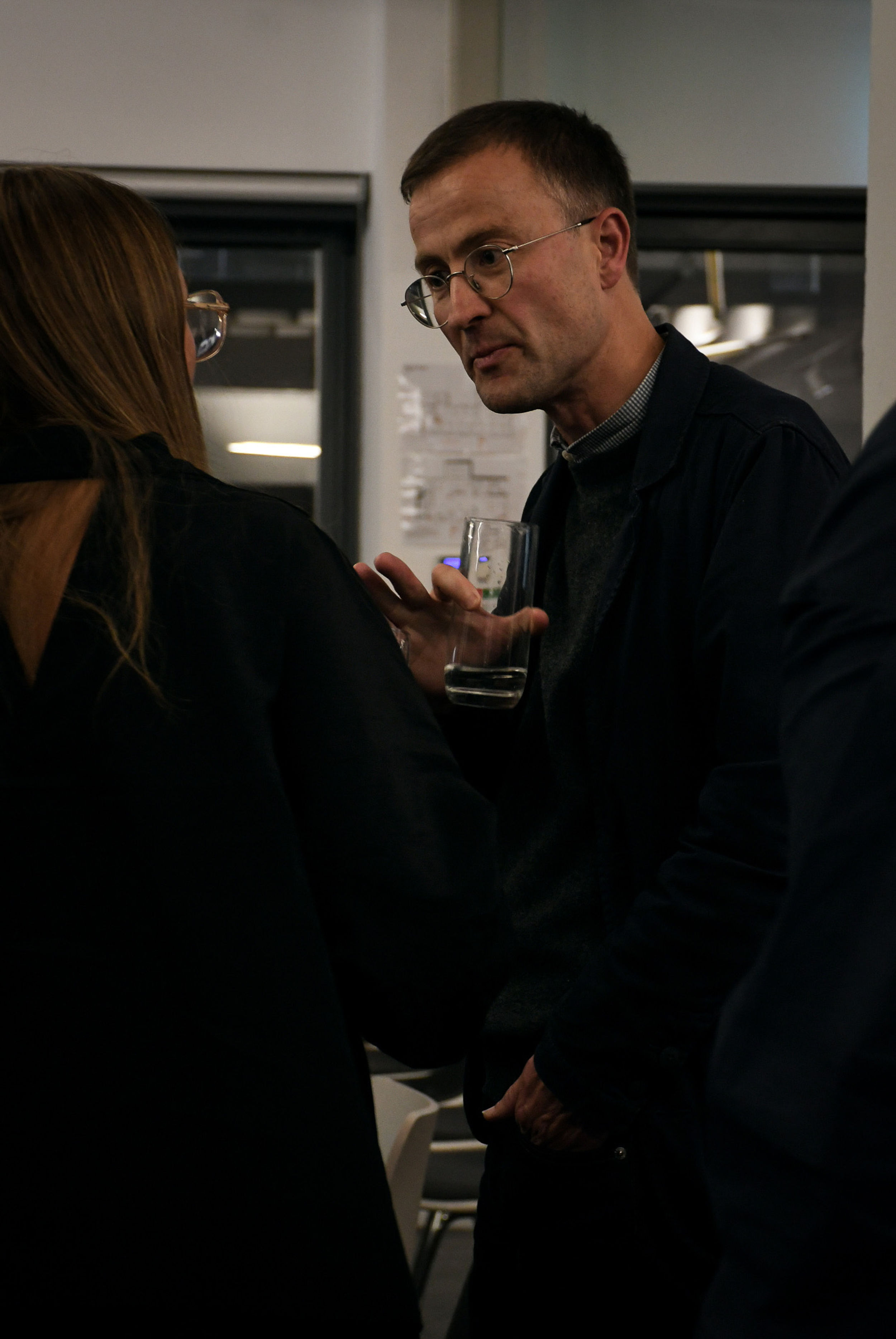 A man wearing glasses holding a glass and talking to a woman.
