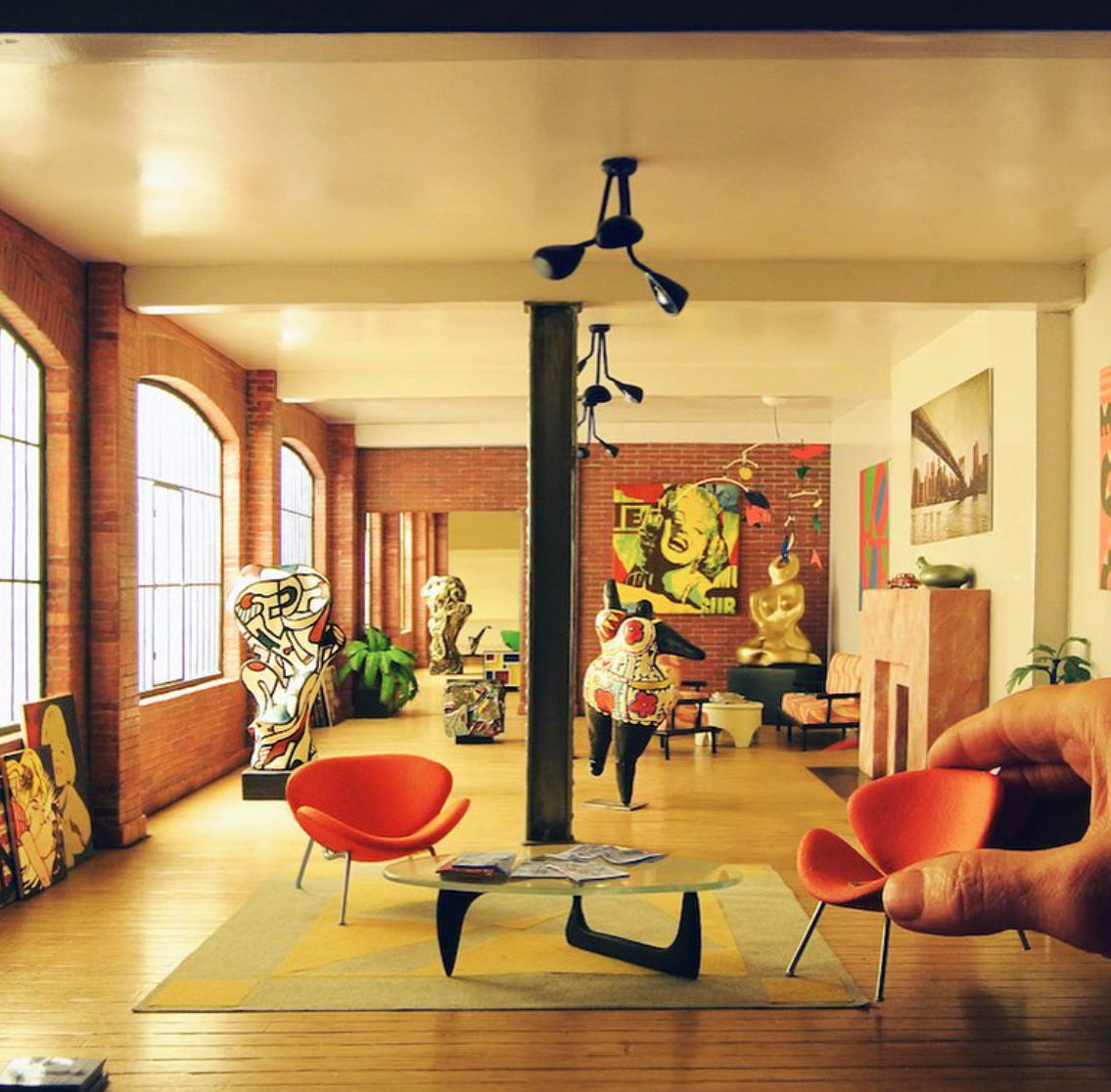 Highly detailed miniature model of a modern art loft with mid-century furniture, pop art sculptures, and bright natural lighting. A human hand adjusts a small orange chair, emphasizing the scale and craftsmanship of the scene.
