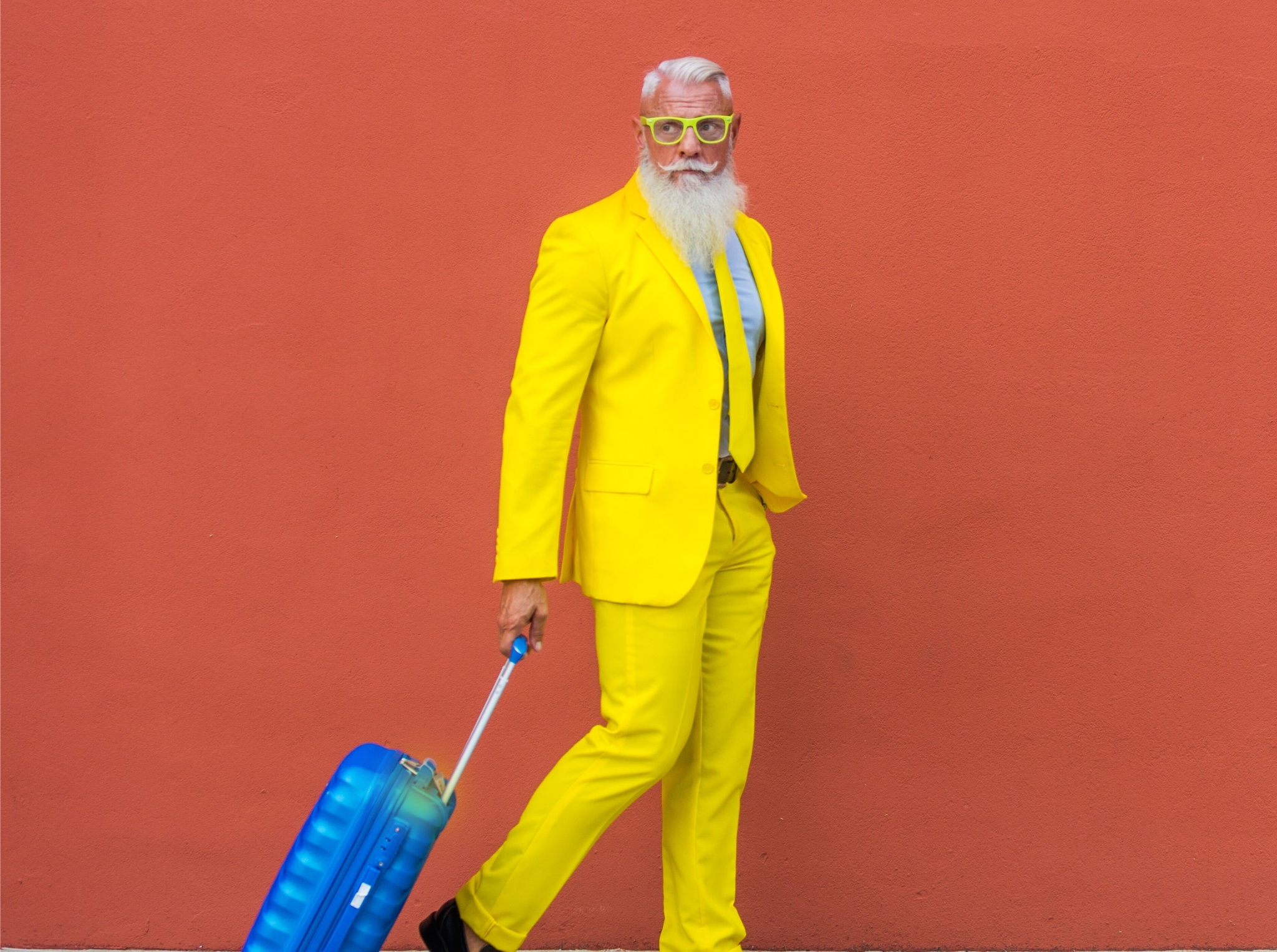 Confident older man in a vibrant yellow suit and glasses pulling a bright blue suitcase against a coral wall—expressing bold, unapologetic travel style for the ‘Planned with Personality’ theme.