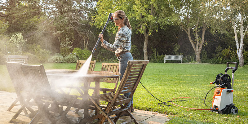 Woman cleaning with the High-pressure cleaner RE 130 PLUS the wooden Garden furniture