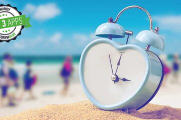 Urlaubscountdown: Herzförmige Uhr auf Sand, im Hintergrund Meer