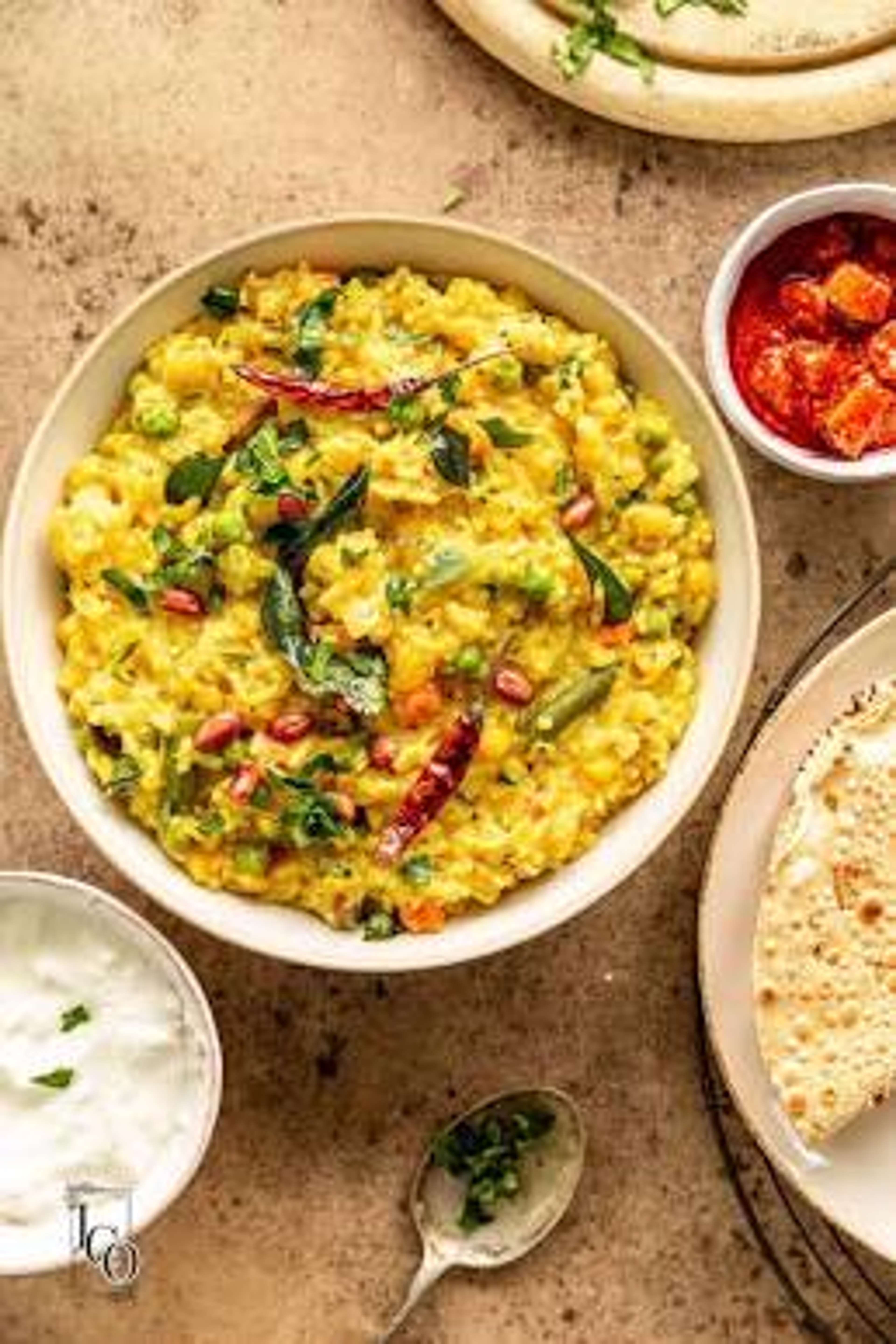 खिचड़ी से भरा एक कटोरा, जिस पर करी पत्ता, मूँगफली और धनिया सजा है; आसपास दही, अचार और पापड़ रखे हैं।
